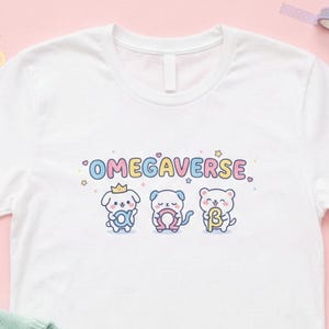 Pode incluir: Camiseta branca com a palavra "OMEGAVERSE" acima de três personagens de animais de desenho animado. Os personagens são um cachorro, um gato e um urso, cada um com um símbolo diferente. A camiseta está em uma superfície rosa com um suéter azul claro e um ursinho branco.