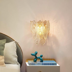 May include: A decorative wall sconce with a clear, ornate glass design. The sconce has multiple layers of glass, creating a cascading effect. A teal balloon dog figurine and a book titled "Monet" are on a nightstand below the sconce.