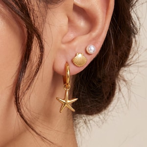May include: Gold-toned earrings featuring a starfish charm, a seashell stud, and a pearl stud. The hoop earrings have a starfish pendant. The seashell stud is detailed with ridges. The pearl stud is round and white. The earrings are worn on an ear.