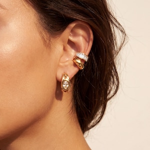 Puede incluir: Juego de joyas doradas que presenta un par de pendientes de aro con piedras claras de forma ovalada, un brazalete de oreja de doble banda y un brazalete de oreja único con piedras claras rectangulares. Las joyas se lucen en la oreja.