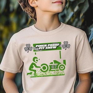 May include: A beige t-shirt featuring a green graphic of a race car and the words "Pinch Proof Pit Crew." The design includes a checkered flag and shamrocks. The number "67" is on the race car.