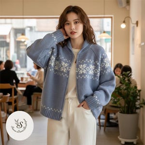 Könnte beinhalten: Hellblauer Reißverschluss-Cardigan mit weißen Schneeflockenmustern. Der Cardigan hat einen gerippten Kragen, Bündchen und Saum. Das Model trägt den Cardigan mit einer weißen Hose. Der Hintergrund zeigt ein Café.