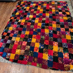 May include: A rectangular quilt constructed from a grid of small, multi-colored fabric squares. The patchwork features a variety of textures and patterns in shades of red, yellow, blue, purple, orange, and black, creating a vibrant, dense mosaic.