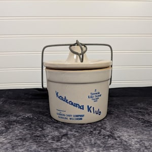 Puede incluir: Una vasija de cerámica beige con tapa a juego y un asa de alambre de metal gris. El frente presenta texto azul que dice Kaukauna Klub, It Spreads Like Butter Copyright 1933 y Manufactured by Kaukauna Dairy Company Kaukauna, Wisconsin.