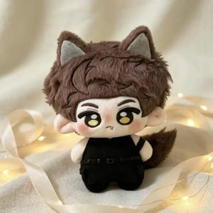 May include: A plush doll with brown fur hair, pointed animal ears, and a fluffy tail. The doll has large embroidered eyes, a small mouth, and pink cheeks. It wears a black sleeveless top with a belt detail around the waist and small ears.
