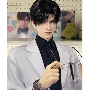 Può includere: Una bambola con i capelli neri, che indossa un blazer bianco sopra una camicia nera e un orologio nero. La bambola tiene in mano occhiali e una penna. Sullo sfondo, ci sono immagini della bambola e di una pera di peluche.