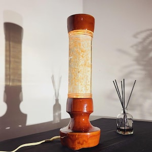 Könnte beinhalten: Eine braune Keramik-Tischlampe mit einem zylindrischen Glaskern, der mit einem hellfarbigen Material gefüllt ist. Die Lampe hat einen abgerundeten Sockel und eine abgerundete Oberseite, und ein Netzkabel erstreckt sich von der Basis. Eine kleine Glasflasche mit Schilf steht neben der Lampe.