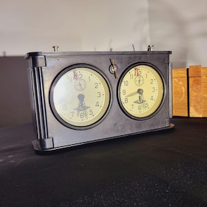 May include: A vintage black chess clock with two circular clock faces. Each face has a cream-coloured background with black numerals and hands. The clock is made of metal and has a rectangular shape.