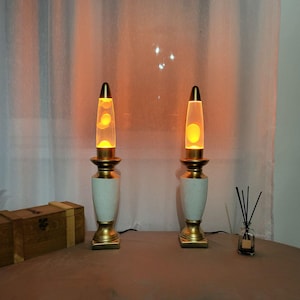 Puede incluir: Dos lámparas de lava retro con bases y tapas doradas, secciones medias blancas y doradas, y tubos de vidrio transparente llenos de lava amarilla. Las lámparas están iluminadas, proyectando un brillo cálido. También se ven una caja de madera y un difusor de caña.