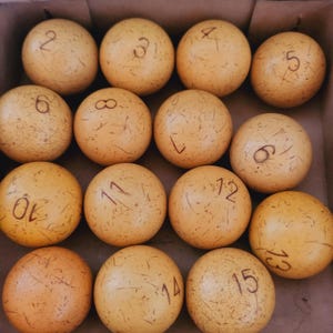 Puede incluir: Una colección de quince bolas de billar esféricas dispuestas en una cuadrícula. Cada bola es de color tostado claro con una textura desgastada y rayada, y presenta un número marrón oscuro escrito a mano del dos al quince, con una bola en blanco.