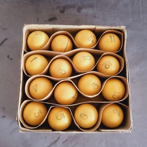 Puede incluir: Una caja de cartón cuadrada que contiene dieciséis esferas de color amarillo mate dispuestas en una cuadrícula de cuatro por cuatro. Cada esfera está marcada con un número negro distinto del uno al dieciséis, separadas por divisores.