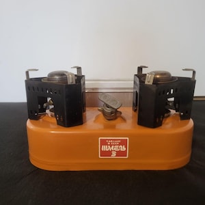 Op de afbeelding: Een oranje kampeerkooktoestel met twee branders, twee zwarte metalen brandersteunen en een centrale tankdop. Een rood rechthoekig label aan de voorzijde bevat de witte tekst "СДЕЛАНО В СССР ШМЕЛЬ 3" in het Cyrillisch schrift.