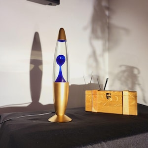 May include: A retro lava lamp with a gold base and cap, and a clear glass cylinder containing blue liquid. The lamp sits on a black surface next to a wooden box with antennas. The lamp's shadow is visible on the wall.