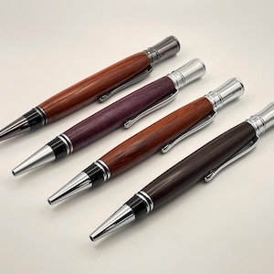 Puede incluir: Cuatro bolígrafos de madera en varios tonos de marrón y morado, con detalles metálicos plateados. Los bolígrafos tienen un aspecto elegante y pulido, con un clip y una punta. Los bolígrafos están dispuestos en diagonal sobre un fondo claro.