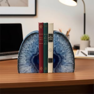 May include: Blue agate bookends holding a row of books with red, green, and white spines. The bookends have a polished, crystalline appearance. The books have gold lettering and are on a wooden desk.