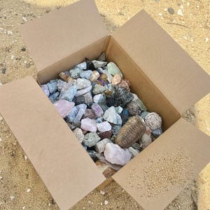 May include: An open cardboard box filled with a variety of colorful rocks and minerals. The collection includes rose quartz, amethyst, and other stones in shades of pink, purple, green, and gray. The box is sitting on a sandy surface.