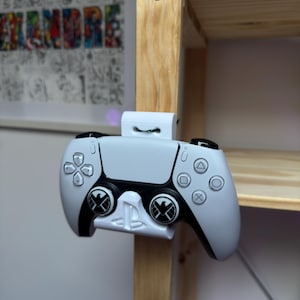 Op de afbeelding: Een wit en zwarte PlayStation 5-controller met een witte montagebeugel, bevestigd aan een houten plank. De controller heeft zwarte accenten en het PlayStation-logo op de knoppen. De achtergrond bevat een ingelijst kunstwerk.