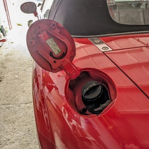 May include: Open red fuel door of a convertible car. The fuel cap is black and visible inside the opening. The car's body is a bright red, with a black convertible top in the background.