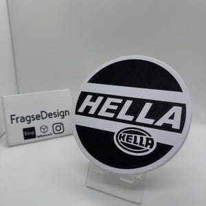 Peut inclure: Un logo HELLA rond, noir et blanc, présenté sur un support transparent. Le logo affiche le mot "HELLA" en blanc, avec un petit logo HELLA en dessous. Un petit panneau blanc avec "Fragsedesign" et des icônes de médias sociaux est visible.