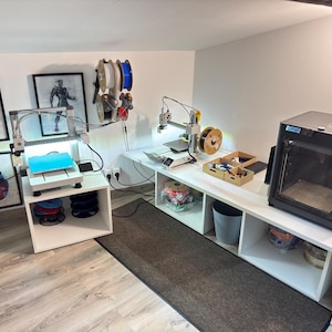 Peut inclure: Un espace de travail avec deux imprimantes 3D à cadre ouvert sur des étagères blanches, une imprimante 3D fermée noire et des bobines de filament fixées au mur. L'espace comprend des affiches de super-héros et des bacs de rangement.