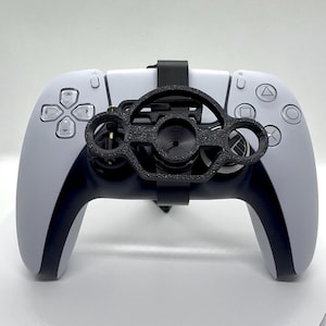 Op de afbeelding: Een wit en zwarte gamecontroller met een zwart accessoire bevestigd. De controller heeft een symmetrisch ontwerp met diverse knoppen en richtingspads. Het accessoire lijkt een telefoonhouder te zijn.