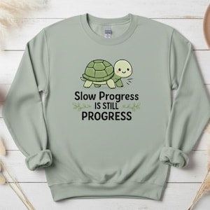 May include: Sage green sweatshirt with a cartoon turtle graphic and the text "Slow Progress is Still Progress." The turtle is green and white. A small potted succulent and a tan hat are in the background.