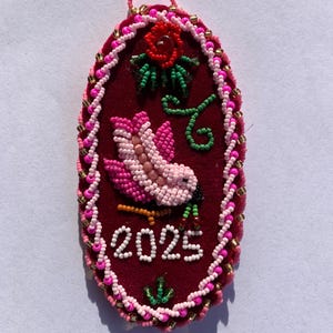 May include: A burgundy beaded ornament with a pink and white bird design, a red rose, and the year 2025. The oval-shaped ornament is trimmed with pink and gold beads and has a pink loop for hanging.