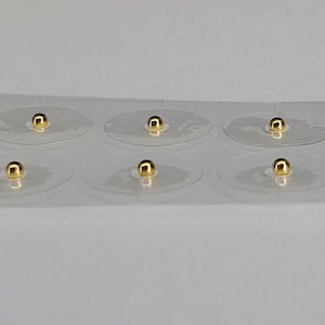 May include: A strip of clear, adhesive dots, each with a small, shiny gold bead in the center. The beads appear to be spherical and reflect light, creating a bright, metallic appearance. The dots are evenly spaced.