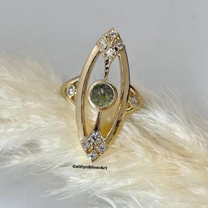 May include: A gold ring with a central green stone and diamond detailing. The ring has an elongated, openwork design with a central oval green stone and diamond clusters at the top and bottom. The ring is set against a soft, white background.