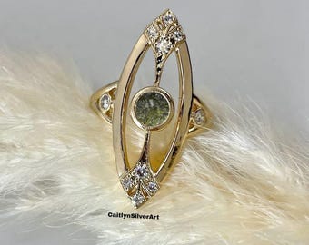 May include: A gold-toned ring with a central green stone and diamond accents. The ring features an elongated, openwork design with a central oval green stone and diamond clusters at the top and bottom. The ring is set against a soft, white background.