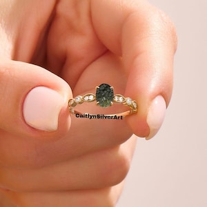 Op de afbeelding: Een gouden ring met een ovale, donkergroene edelsteen. De band is versierd met kleine, heldere edelstenen en een decoratief patroon. De ring wordt tegen een zachte roze achtergrond gehouden. De tekst "CaitlynSilverArt" is zichtbaar.