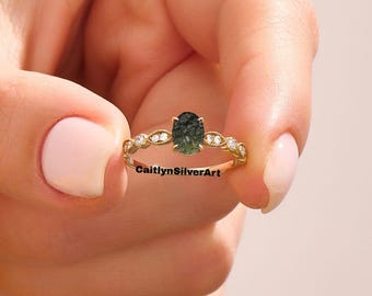 May include: A gold ring featuring an oval-cut, dark green gemstone. The band is adorned with small, clear gemstones and a decorative pattern. The ring is held up against a soft pink background. The text "CaitlynSilverArt" is visible.