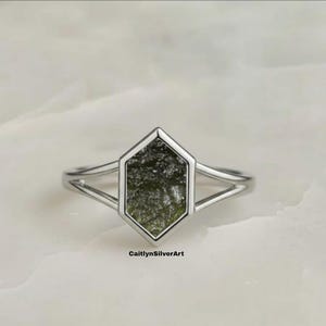 Op de afbeelding: Een zilveren ring met een zeshoekige groene steen. De ring heeft een gesplitst bandontwerp. De steen heeft een textuur. De ring is geplaatst op een wit marmeren oppervlak. De tekst "CaitlynSilverArt" is zichtbaar.