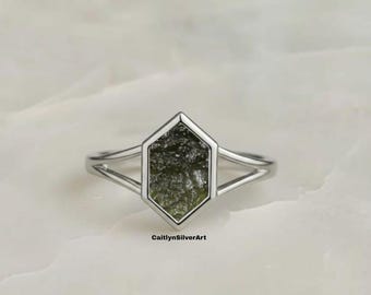 May include: A silver ring featuring a hexagonal green stone. The ring has a split band design. The stone has a textured appearance. The ring is set against a white marble surface. The text "CaitlynSilverArt" is visible.