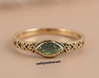 May include: A gold-colored ring with an oval, green stone at the center. The band features an ornate, filigree design. The text "caitlynsilverart" is printed at the bottom of the image.