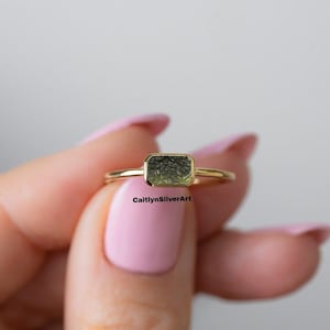 Könnte beinhalten: Ein goldener Ring mit einem rechteckigen, olivgrünen Edelstein. Der Ring hat einen dünnen, polierten Ring und eine Lünettenfassung. Der Text "CaitlynSilverArt" ist auf dem Bild sichtbar. Dieser Ring ist ein Schmuckstück.