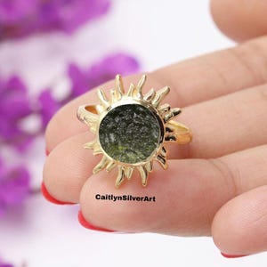 Könnte beinhalten: Ein goldfarbener Ring mit Sonnenstrahlen-Design. Der Ring hat einen zentralen, runden, dunkelgrünen Stein. Der Ring wird in einer Hand mit rotem Nagellack gehalten. Der Text "CaitlynSilverArt" ist sichtbar.