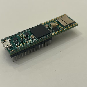 May include: A small, green circuit board with various electronic components, including a USB port, a black rectangular chip, and a slot for a micro SD card. The board has numerous small holes and metal pins along the bottom edge.