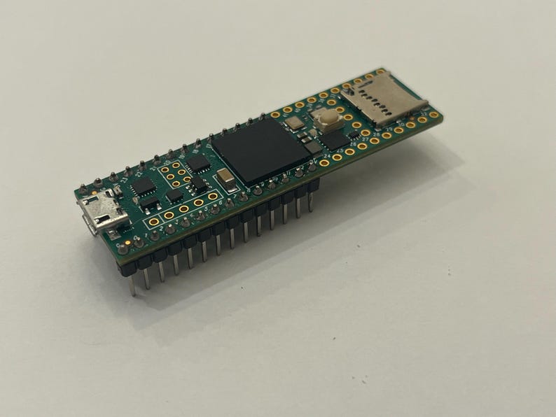 May include: A small, green circuit board with various electronic components, including a USB port, a black rectangular chip, and a slot for a micro SD card. The board has numerous small holes and metal pins along the bottom edge.