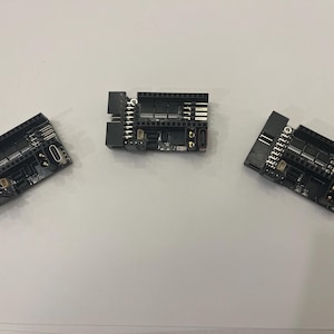May include: Three black electronic circuit boards with various components, including pins, ports, and chips, are arranged on a white surface. The boards have a rectangular shape with multiple connectors and are likely used for electronics projects.