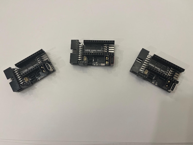 May include: Three black electronic circuit boards with various components, including pins, ports, and chips, are arranged on a white surface. The boards have a rectangular shape with multiple connectors and are likely used for electronics projects.