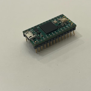 May include: A small, green circuit board with various electronic components, including a black square chip, a USB port, and numerous gold-colored pins. The board is designed for electronics projects and prototyping.