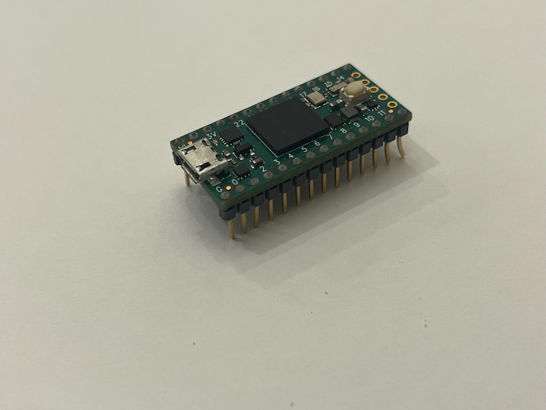 May include: A small, green circuit board with various electronic components, including a black square chip, a USB port, and numerous gold-colored pins. The board is designed for electronics projects and prototyping.