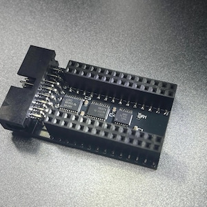 May include: A black circuit board with multiple connectors and integrated circuits. The board has rows of pins and several small chips labeled with text. The components are arranged on a dark green surface.