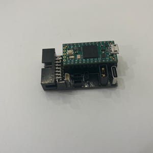 May include: A small, green circuit board with various electronic components, including a USB port and multiple pins. The board is connected to a black base with additional ports and connectors. The overall design suggests a microcontroller or development board.
