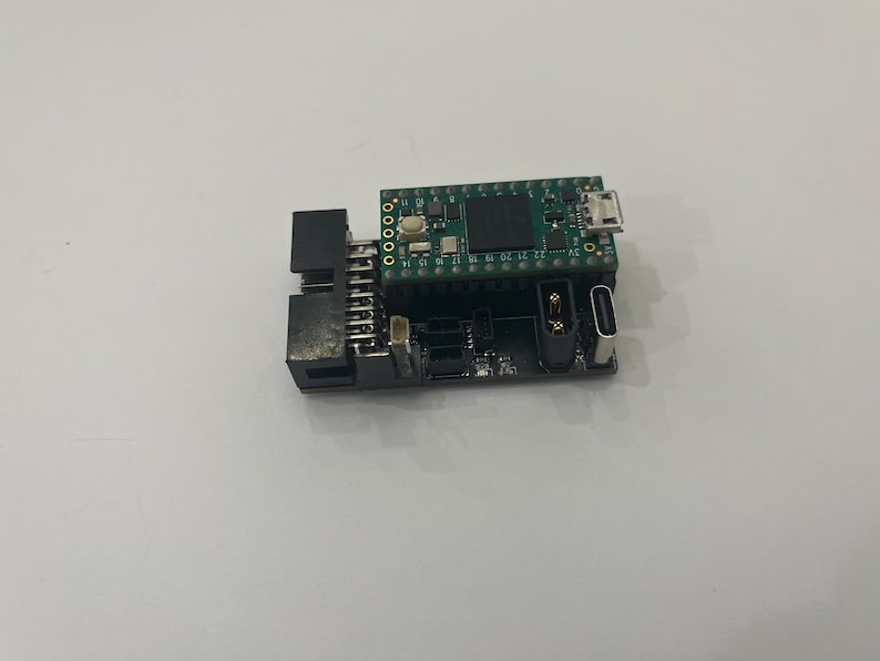 May include: A small, green circuit board with various electronic components, including a USB port and multiple pins. The board is connected to a black base with additional ports and connectors. The overall design suggests a microcontroller or development board.