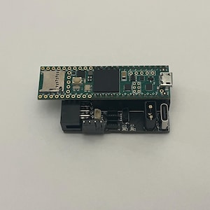 May include: A small, dark green circuit board with various electronic components, including a micro SD card slot and USB port, is stacked on top of a black circuit board. The boards are set against a white background.
