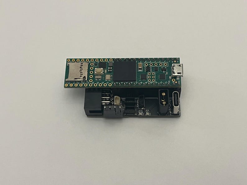 May include: A small, dark green circuit board with various electronic components, including a micro SD card slot and USB port, is stacked on top of a black circuit board. The boards are set against a white background.