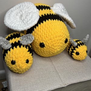 May include: Three handmade crochet bees in varying sizes. The bees are yellow with black stripes and have white or grey wings. The largest bee is in the centre, with two smaller bees on either side. The bees have black safety eyes.