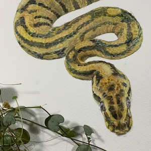 Może przedstawiać: Akwarela przedstawiająca węża z żółtymi, brązowymi i czarnymi oznaczeniami. Wąż jest zwinięty na białym tle. Zielone, liściaste pnącza znajdują się na pierwszym planie. Głowa węża znajduje się w prawym dolnym rogu.